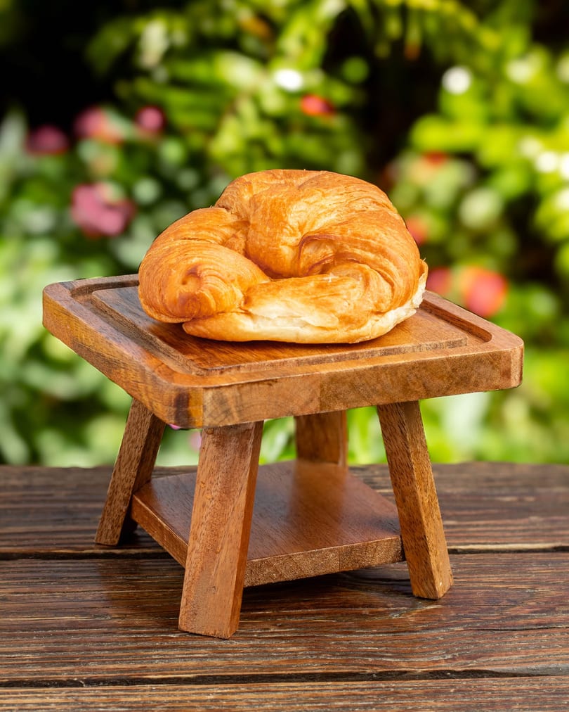 Golden-brown croissant on a small wooden stand, with a blurred green garden background.