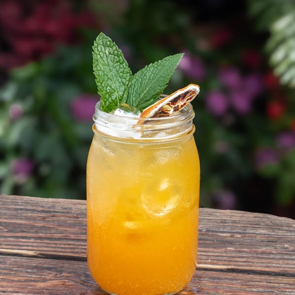 A vibrant orange cocktail in a mason jar, garnished with mint and a dried orange slice, on a wooden table.
