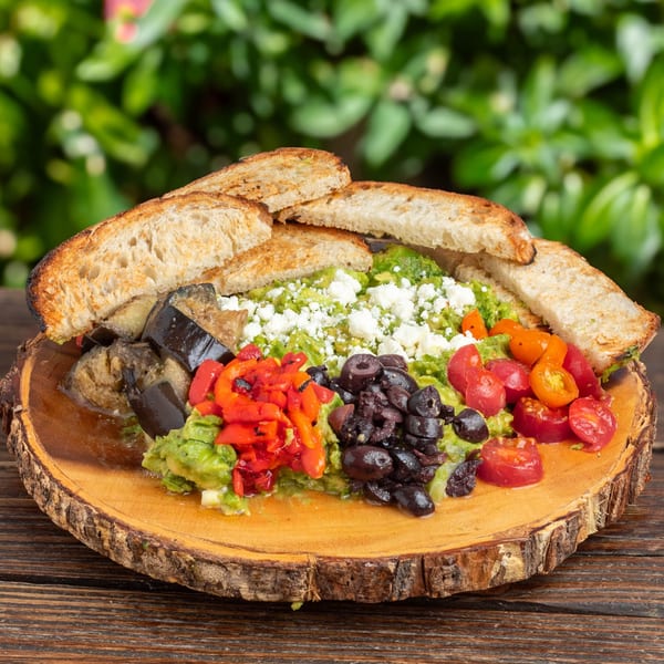 Avocado toast with feta, roasted peppers, eggplant, olives, and tomatoes on a wooden board.
