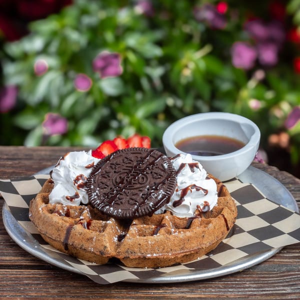 A waffle topped with whipped cream, chocolate syrup, an Oreo cookie, and sliced strawberries.