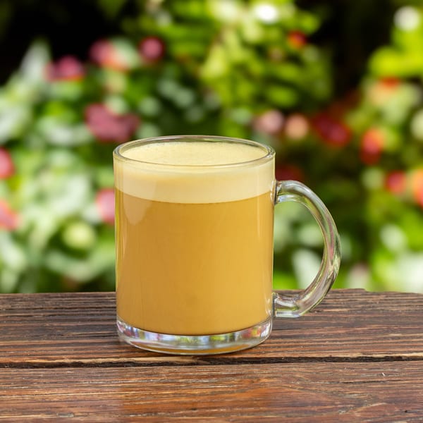 A clear mug of golden frothy beverage sits on a wooden table with a blurred green and pink background.