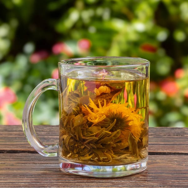 Blooming tea in a clear glass mug on a wooden table with a blurred green and pink background.