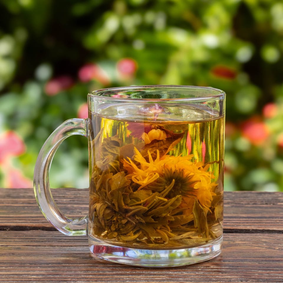 Blooming tea in a clear glass mug on a wooden table with a blurred green and pink background.
