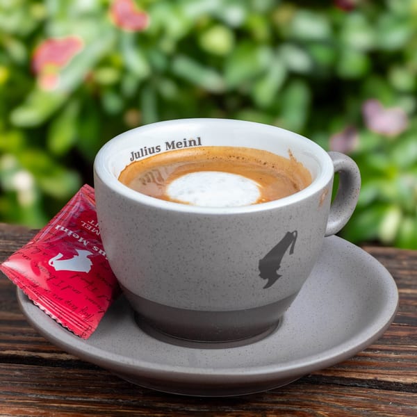 A Julius Meinl coffee cup with foam and a red sugar packet on a wooden table outdoors.