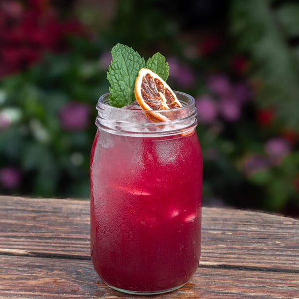 A vibrant red drink in a mason jar, garnished with mint and a dried orange slice, on a wooden table.