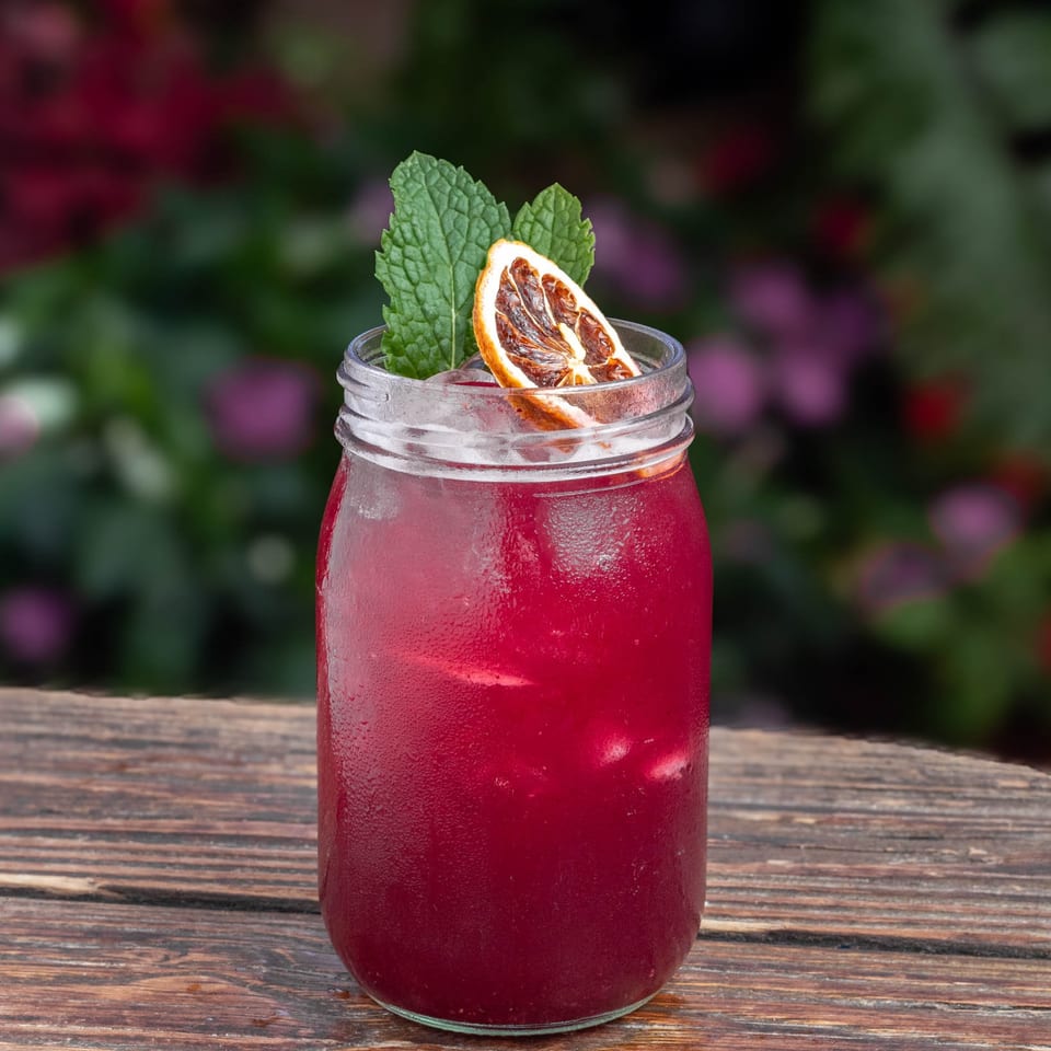 A vibrant red drink in a mason jar, garnished with mint and a dried orange slice, on a wooden table.