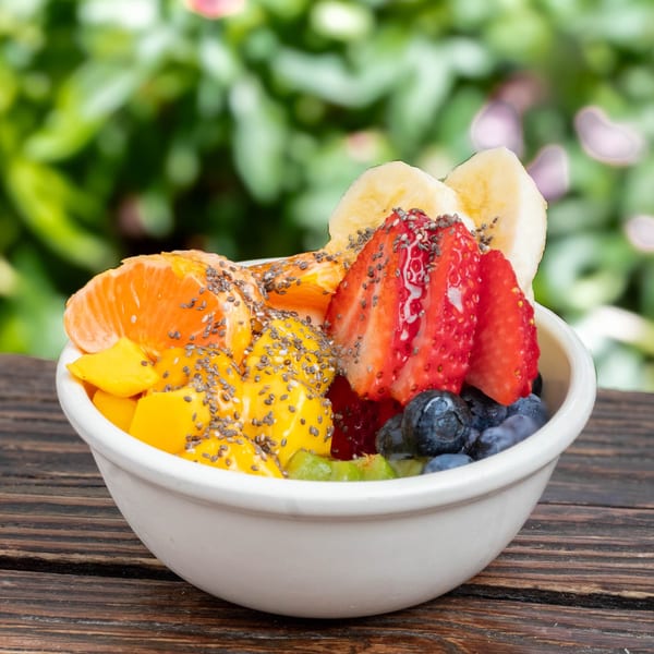A white bowl filled with fresh fruit, including mango, strawberries, blueberries, and bananas, on a wooden table.