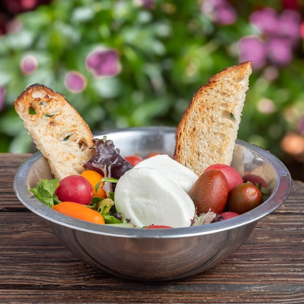 Caprese salad with fresh mozzarella, colorful tomatoes, mixed greens, and toasted bread in a metal bowl.